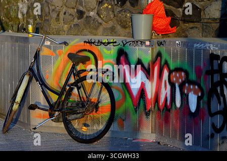 Altstadtrad lehnt sich an eine Graffiti-bedeckte Metallwand neben einem Damm, Amsterdam, Niederlande, warmes Abendlicht mit sichtbarem Eimer. Stockfoto