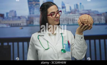 Frau Ärztin mit Hirnmodell an der Strandpromenade mit Stethoskop in Uniform im Freien, selbstbewusst lächelnd. Stockfoto