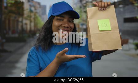 Frau in blauer Uniform hält Papiertüte, lächelt und zeigt mit dem Finger auf den Gegenstand auf der Straße; Kundenzufriedenheit. Stockfoto