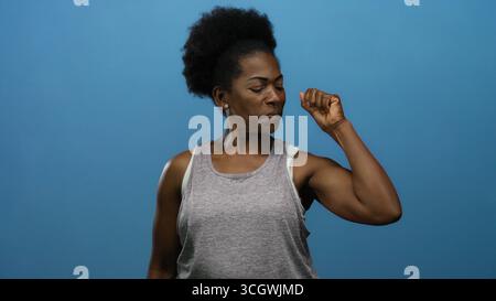 Frau beugt Muskeln in lässiger Kleidung vor isoliertem blauem Hintergrund und zeigt Empowerment und Kraft. Stockfoto