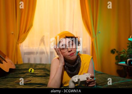 Teenager-Mädchen in entspannter Körperhaltung zu Hause im Abendlicht. Gemütliche Umgebung mit Gitarre und warmen Kissen. Jugendfreiheit und Technik. Mitte Stockfoto