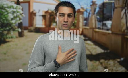 Der junge Mann steht draußen in einer Parklandschaft und drückt seine Kontemplation mit Hand auf Brust aus, vor einem Hintergrund von Bäumen und einem sonnigen Tag. Stockfoto