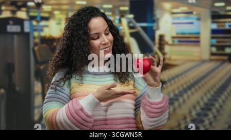 Die lächelnde Frau hält das Herz, während sie im Fitnessstudio mit einem farbenfrohen Pullover die Daumen nach oben drückt und im Fitnesscenter positiver wirkt. Stockfoto