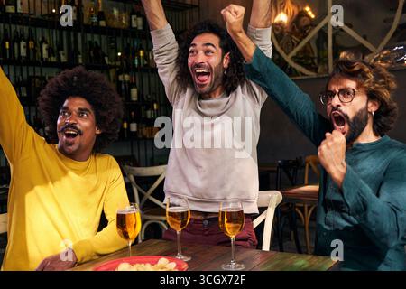Multirassische Gruppe von männlichen Freunden, die Fußballspiele an der Bar anschauen. Feiern Sie eine Partitur mit den Fäusten Stockfoto