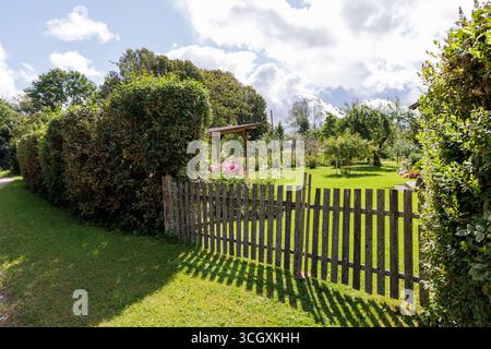 Ein alter Holzzaun wirft Schatten auf einen grünen Rasen und führt in einen üppigen Garten mit Büschen und blühenden Pflanzen. Stockfoto