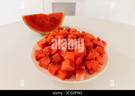 Saftige, lebendige Wassermelonenwürfel sind ordentlich auf einer weißen Platte angeordnet, begleitet von einer halben Wassermelone im Hintergrund. Diese erfrischende Frucht ist Stockfoto