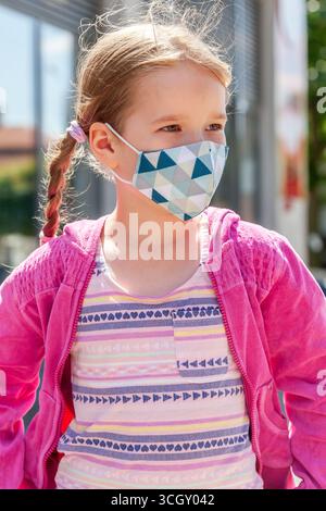 Schulalter Mädchen trägt moderne Art Schutzmaske im Freien an sonnigem Tag, stilvolle rosa Jacke, geflochtene Haare, Pandemie Virus Sicherheitsvorkehrungen Stockfoto