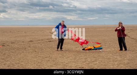 St Annes, Lancashire, Großbritannien – 30. August 2025: Kite Festival. Zwei Personen bereiten sich darauf vor, an einem Sandstrand unter bewölktem Himmel Drachen zu fliegen. Stockfoto
