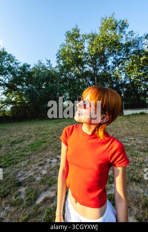 Junge Rothaarige Frau mit Sonnenbrille, die den Sonnenuntergang in einem Park genießt Stockfoto