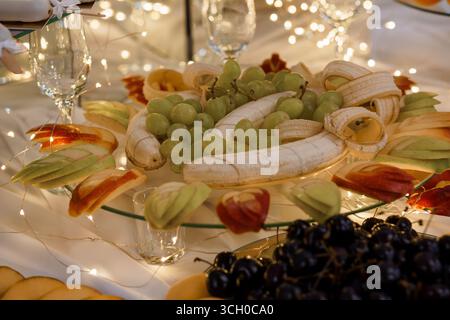Ein lebendiges Bild über dem Kopf mit einer wunderschön angeordneten Obstplatte, voll mit geschnittenen Äpfeln, Bananen, grünen Trauben und dunklen Kirschen, Akzenten Stockfoto