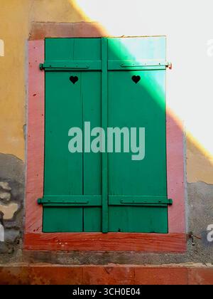 Fenster mit grünen Fensterläden und Herzdetails auf einer korallenroten und gelben Hauswand mit Natursteinen aus dem Elsass, Frankreich Stockfoto