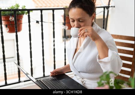 Eine Frau sitzt auf einem Balkon, trägt einen Bademantel, schlürft Kaffee und stöbert auf ihrem Laptop. Die Szene fängt das Wesen eines ruhigen und produktiven Motivs ein Stockfoto