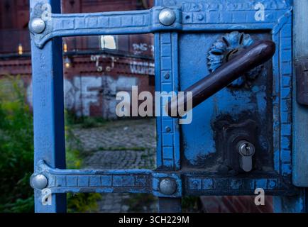 Detaillierte Ansicht des alten blauen Metalltors mit kunstvollem Griff und Schloss, Backsteinarchitektur im Hintergrund leicht verschwimmt. Stockfoto