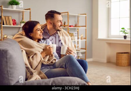Liebevolles, liebevolles Paar, das in einem modernen Wohnzimmer sitzt und mit einer warmen Decke umhüllt ist, fröhlich romantisch Stockfoto