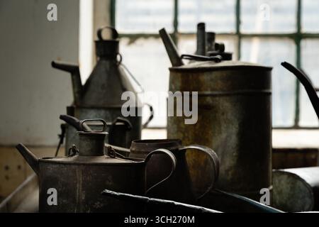 Alte Ölkannen und -Krüge aus Metall auf einer Werkbank in einer industriellen Werkstatt, beleuchtet von Fensterlicht, zeigt Rost und Patina. Stockfoto