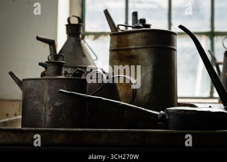 Alte Ölkannen und -Krüge aus Metall auf einer Werkbank in einer industriellen Werkstatt, beleuchtet von Fensterlicht, zeigt Rost und Patina. Stockfoto