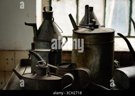 Alte Ölkannen und -Krüge aus Metall auf einer Werkbank in einer industriellen Werkstatt, beleuchtet von Fensterlicht, zeigt Rost und Patina. Stockfoto