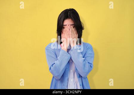 Eine junge Frau in einem blauen Cardigan wirkt nachdenklich, schlecht gelaunt und stressig vor einem leuchtend gelben Hintergrund Stockfoto