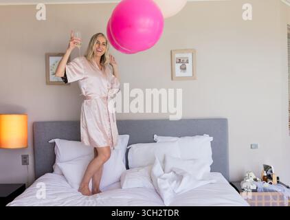 Junge Frau, die im modernen Schlafzimmer auf dem Bett steht und mit pinken Ballons und Weinglas feiert Stockfoto