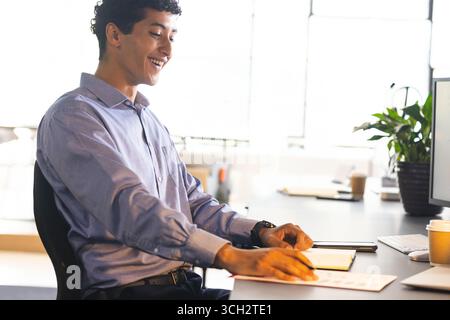 Mann, der Dokumente am Schreibtisch vor großen Fenstern liest, mit Monitor und Kaffeetasse Stockfoto