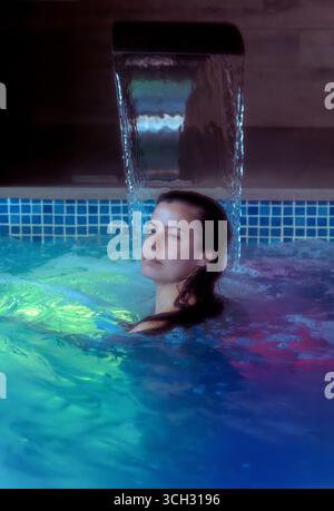 Eine schöne junge Frau genießt entspannende und friedliche Hydrotherapie in einem Spa-Pool mit farbigen Lichtern im Wasser. Stockfoto