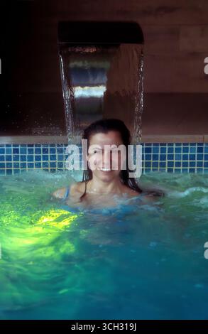 Eine schöne, lächelnde junge Frau genießt eine entspannende und friedliche Hydrotherapie-Sitzung unter einem Wasserfall in einem Spa-Pool mit farbigen Lichtern und sprudelnden Lichtern Stockfoto