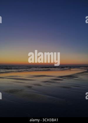 Ein Sonnenuntergang bietet ein majestätisches und farbenfrohes Spektakel am Sandstrand von Chipiona mit Menschen, die umherschlendern, in einer Küstenstadt in Andalusien, Spanien Stockfoto