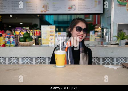 Ho-Chi-Minh-Stadt, Vietnam - 10. Mai 2025: Eine junge Frau mit Sonnenbrille und Schal lächelt, während sie an einem Tisch in einem Flughafencafé mit einem Drink sitzt. Stockfoto
