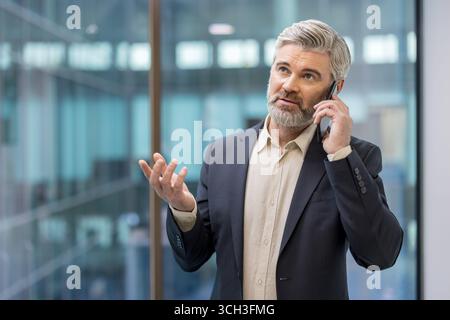 Ein selbstbewusster Mann in einem Anzug, der einen Anruf führt und in einem modernen Büroumfeld steht. Stockfoto