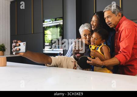 Mehrgenerationenfamilie macht Selfie in der Küche, feiert mit Trophäe und lächelt zu Hause Stockfoto