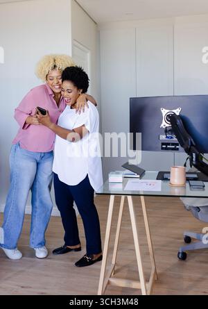 Freunde, die sich umarmen und lachen, während sie im modernen Büro auf das Smartphone schauen Stockfoto