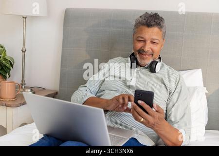 Seniorenmann, der sich mit Smartphone und Laptop im Bett entspannt und Kopfhörer trägt Stockfoto