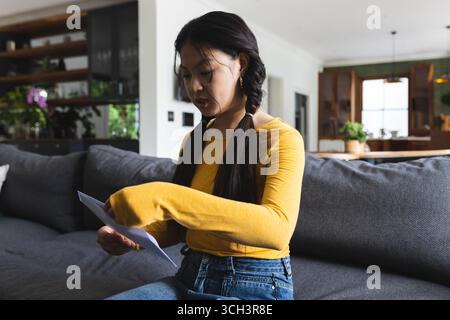 Asiatische Frau mit weißem Umschlag auf grauem Sofa mit Topfpflanzen im modernen Wohnbereich Stockfoto