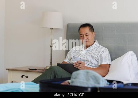 Asiatischer Mann, der sich zu Hause mit Smartphone und Tablet im Bett entspannt Stockfoto