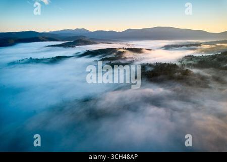 Atemberaubender Blick aus der Vogelperspektive bei Sonnenaufgang mit einem riesigen Wolkenmeer, das die Berglandschaft umhüllt. Subtile Farbtöne der aufgehenden Sonne färben den Himmel in sanften Pastelltönen und schaffen eine ruhige und ätherische Szene. Stockfoto