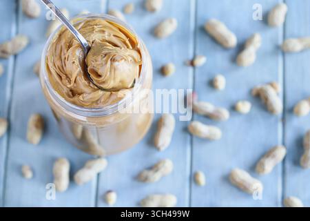 Erdnussbutter wird mit einem Löffel aus einem Plastikbehälter über einem blauen Tisch mit verstreuten Nüssen in der Schale aufgeschlüsselt. Selektiver Fokus mit Unschärfe Stockfoto