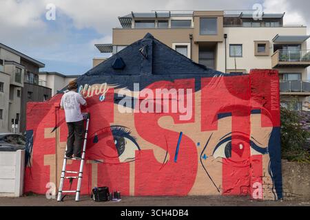 Southend on Sea, Großbritannien. 31. August 2025. Ein Straßenkünstler malt ein großes Wandbild mit einem stilisierten Gesicht und dem Wort „SCHNELL“ an einer Wand in Southend-on-Sea. Die Szene fängt den Akt der Schöpfung ein, mit dem Künstler auf einer Leiter und modernen Wohnungen im Hintergrund. Der dritte Tag von City Jam. Viele Künstler arbeiten noch an ihren Wandmalereien. 2025 ist das vierte Jahr, in dem die Veranstaltung stattfindet. Penelope Barritt/Alamy Live News Stockfoto