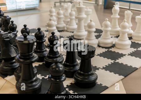 Große schwarz-weiße Schachfiguren sind auf einem karierten Brett in einem Gemeindezentrum angeordnet. Die Spieler treffen sich und bereiten sich auf ein strategisches Spiel vor Stockfoto