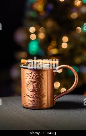 Festlicher Weihnachtscocktail, serviert in einem Tito’s Handmade Vodka Kupferbecher mit Eis und einer Zimtstange, vor einem warmen Weihnachtshintergrund Stockfoto