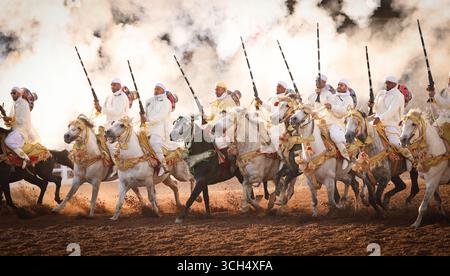 Eine Geschichte von Mut, Einheit und Erbe in einem einzigen Bild. Reiter in bunten marokkanischen Kostümen galoppieren in einer Fantasia-Show auf arabischen Pferden Stockfoto
