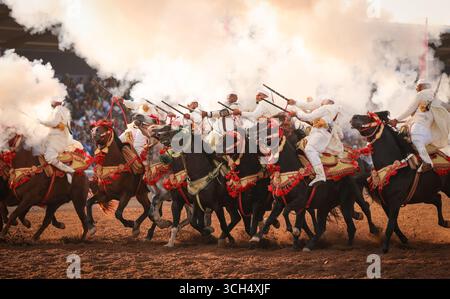 Eine Geschichte von Mut, Einheit und Erbe in einem einzigen Bild. Reiter in bunten marokkanischen Kostümen galoppieren in einer Fantasia-Show auf arabischen Pferden Stockfoto