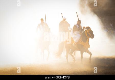 Eine Geschichte von Mut, Einheit und Erbe in einem einzigen Bild. Reiter in bunten marokkanischen Kostümen galoppieren in einer Fantasia-Show auf arabischen Pferden Stockfoto