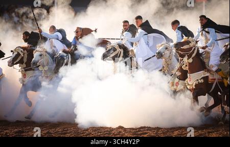 Eine Geschichte von Mut, Einheit und Erbe in einem einzigen Bild. Reiter in bunten marokkanischen Kostümen galoppieren in einer Fantasia-Show auf arabischen Pferden Stockfoto