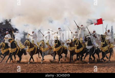 Eine Geschichte von Mut, Einheit und Erbe in einem einzigen Bild. Reiter in bunten marokkanischen Kostümen galoppieren in einer Fantasia-Show auf arabischen Pferden Stockfoto