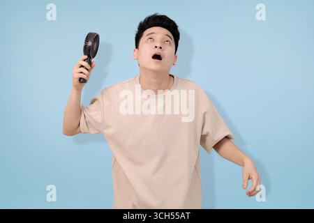 Ein junger Mann, der einen tragbaren Handventilator hält, der Erleichterung vor blauem Hintergrund ausdrückt Stockfoto