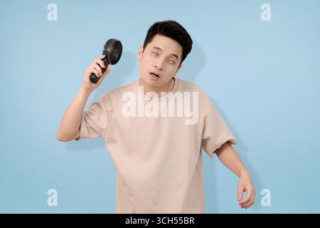 Ein junger Mann, der einen tragbaren Handventilator hält, der Erleichterung vor blauem Hintergrund ausdrückt Stockfoto