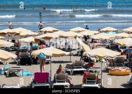 Lebhafte Strandszene in Playa del Inglés, mit Reihen von Sonnenschirmen und Liegestühlen auf dem Sand, Menschen, die sich entspannen und spazieren gehen und Schwimmer im Meer. Stockfoto