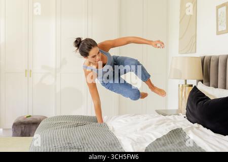 Frau, die über das Bett in einem modernen Schlafzimmer mit weißen Schranktüren und grauer Ottomane springt Stockfoto