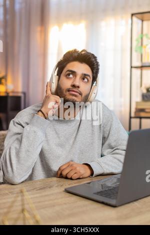 Junger indischer Mann mit schnurlosen Kopfhörern, der am Laptop arbeitet und beim Hörbuch konzentriert wirkt. Der hispanische Kerl sitzt auf dem Sofa am Tisch im Wohnzimmer zu Hause, tief in seine Aufgabe verwickelt. Stockfoto