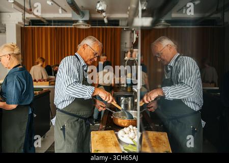 Senior Mann, der während des Kochkurses in der kommerziellen Küche das Essen in der Pfanne brät Stockfoto